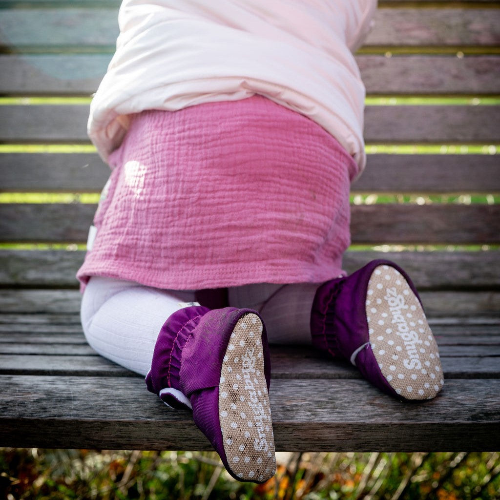 Child in a pink coat wearing Snugabugz plum water-resistant booties that stay on sitting on a park bench
