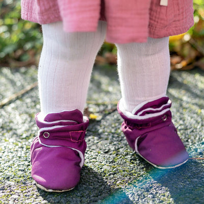 Purple waterproof baby booties that stay on worn by a child on a pavement with a pink skirt and tights.