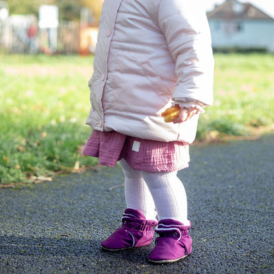 Child wearing a pink coat, pink dress, and purple water-resistant stay on baby booties standing on a path with grass and a playground in the background.