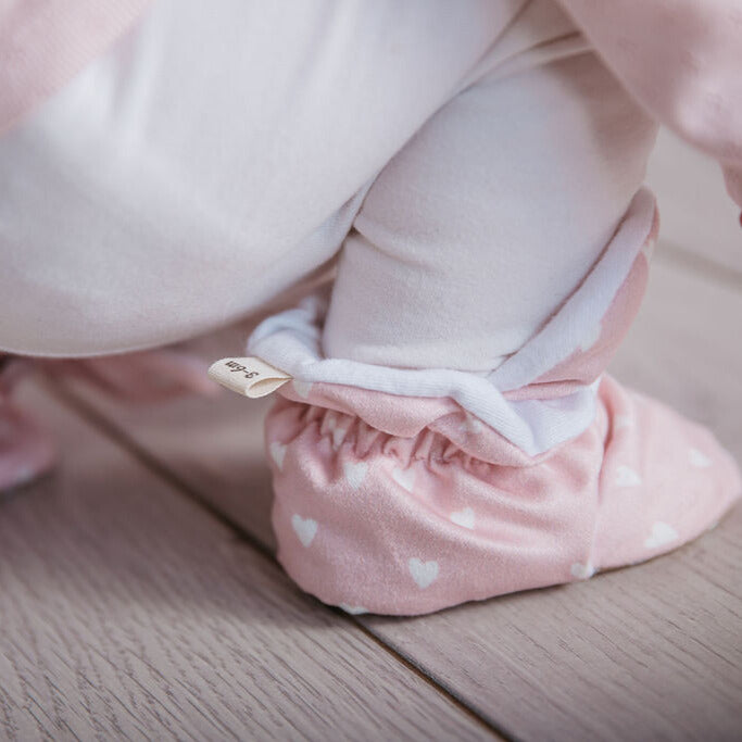 Baby girl squatting wearing a pair of Snugabugz pink stay-on baby booties featuring a white heart print on cotton fabric, with non-slip soles and adjustable three snap closure. Made from organic cotton.