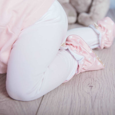 Baby girl crawling wearing a pair of Snugabugz pink baby booties that don't fall off featuring a white heart print on cotton fabric, with non-slip soles and adjustable three snap closure. Pram shoes made from organic cotton. baby slippers with grip soles.